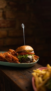 A gourmet burger with layers of beef, lettuce, tomato, and cheese is placed on a wooden table. A skewer holds the burger together. To the side, there are sweet potato fries, and another plate with regular fries is blurred in the foreground.