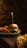 A gourmet burger with layers of beef, lettuce, tomato, and cheese is placed on a wooden table. A skewer holds the burger together. To the side, there are sweet potato fries, and another plate with regular fries is blurred in the foreground.