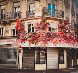 A street corner features a charming caf&eacute; with a facade covered in lush, vibrant flowers. The building showcases classic Parisian architecture, complete with ornate black iron balconies and cream-colored stonework. The red and pink floral arrangements cascade down the side, adding a burst of color to the urban scene.