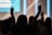 Silhouettes of people with raised arms, likely in a concert or worship setting. The background is blurred with bright vertical lights.
