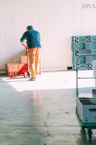 A person wearing a dark green jacket and brown pants is operating a red pallet jack with several cardboard boxes stacked on it. There are plastic storage bins leaning against the wall to the right, and additional items are placed on a cart in the foreground. The setting appears to be a warehouse or storage facility with concrete flooring and a white paneled wall.