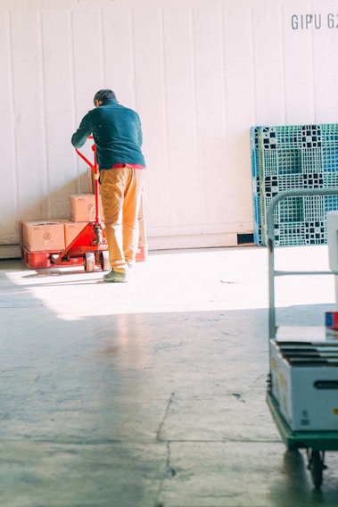 A person wearing a dark green jacket and brown pants is operating a red pallet jack with several cardboard boxes stacked on it. There are plastic storage bins leaning against the wall to the right, and additional items are placed on a cart in the foreground. The setting appears to be a warehouse or storage facility with concrete flooring and a white paneled wall.