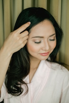 A woman with long, dark hair and a serene expression gently touches her head while smiling softly. She wears a light-colored robe against a neutral background.