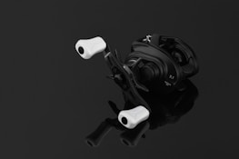 A sleek, modern fishing reel with a black body and white handles, positioned on a reflective black surface. The reel has inscriptions indicating specifications.
