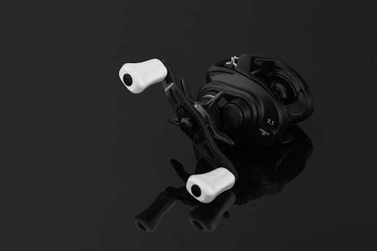 A sleek, modern fishing reel with a black body and white handles, positioned on a reflective black surface. The reel has inscriptions indicating specifications.