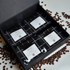 A sleek black box open to reveal an assortment of coffee samples and a small teal coffee scoop inside.