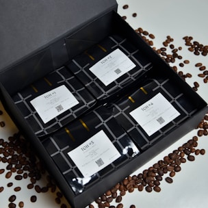 A neatly arranged black gift box containing four packages of coffee, labeled from TOH #1 to TOH #4. Each package has a white label with text and a QR code. Coffee beans are scattered around the box on a white surface.