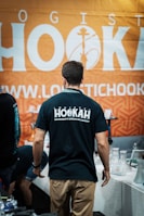 A man in a black T-shirt with 'HOOKAH' written on the back stands facing away from the camera. He is in front of a booth that has various hookah accessories on display. The background features a large orange banner with white text and a hookah logo.