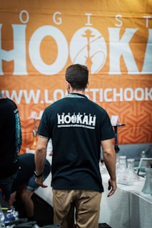 A man in a black T-shirt with 'HOOKAH' written on the back stands facing away from the camera. He is in front of a booth that has various hookah accessories on display. The background features a large orange banner with white text and a hookah logo.