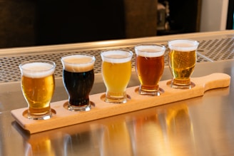 Five small beer glasses filled with different types of beer are arranged in a row on a wooden paddle. Each glass contains a distinct color reflecting various beer styles, from light golden to dark stout. The setting is on a shiny metallic surface, likely a bar counter.