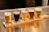 Five small beer glasses filled with different types of beer are arranged in a row on a wooden paddle. Each glass contains a distinct color reflecting various beer styles, from light golden to dark stout. The setting is on a shiny metallic surface, likely a bar counter.