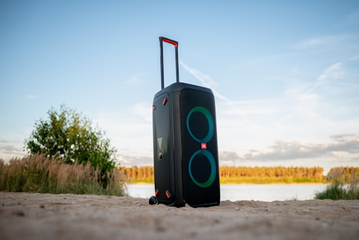 A large portable speaker with a retractable handle and wheels is placed on sandy ground near a body of water, with greenery and a clear sky in the background. The speaker has two circular speaker grills that emit a glow.