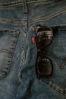 A pair of black sunglasses is resting on the pocket of blue denim jeans, showing the reflection of a window or outdoor scene on the lenses. The brand tag on the jeans is visible.