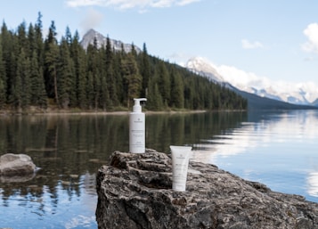 Two skincare products are placed on a large rock by a calm lake, surrounded by dense pine forests and distant mountains. The clear sky above is reflected in the tranquil water.