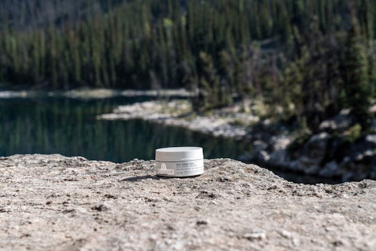 A jar of cream is placed on a large, textured rock with a calm lake and dense forest in the background. The scene suggests a natural, serene outdoor setting.