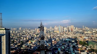 A sprawling urban cityscape with numerous high-rise buildings under a clear blue sky. A prominent telecommunications tower is situated in the middle of the image, surrounded by densely packed buildings. The horizon shows a mix of modern skyscrapers and older structures.