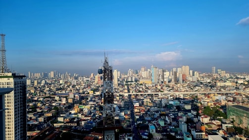 A sprawling urban cityscape with numerous high-rise buildings under a clear blue sky. A prominent telecommunications tower is situated in the middle of the image, surrounded by densely packed buildings. The horizon shows a mix of modern skyscrapers and older structures.