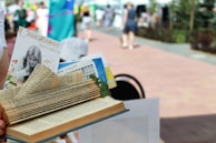 A creatively folded book with various pages tucked and shaped to appear decorative. A pamphlet and other materials are inserted between the pages. The background is a blurred outdoor setting with people walking on a paved path, suggesting a festival or event.