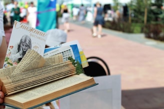 A creatively folded book with various pages tucked and shaped to appear decorative. A pamphlet and other materials are inserted between the pages. The background is a blurred outdoor setting with people walking on a paved path, suggesting a festival or event.