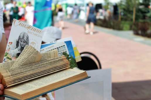 A creatively folded book with various pages tucked and shaped to appear decorative. A pamphlet and other materials are inserted between the pages. The background is a blurred outdoor setting with people walking on a paved path, suggesting a festival or event.