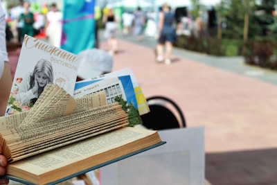 A creatively folded book with various pages tucked and shaped to appear decorative. A pamphlet and other materials are inserted between the pages. The background is a blurred outdoor setting with people walking on a paved path, suggesting a festival or event.