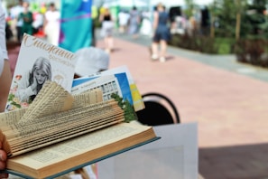 A creatively folded book with various pages tucked and shaped to appear decorative. A pamphlet and other materials are inserted between the pages. The background is a blurred outdoor setting with people walking on a paved path, suggesting a festival or event.