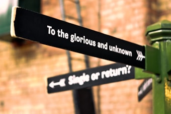 A signpost with multiple black directional signs featuring white text. One sign reads 'To the glorious and unknown' with an arrow, another sign reads 'Single or return?', and a third partially visible sign is present. The background has a blurred brick wall and a vertical metal ladder in focus.
