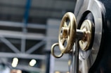A close-up view of a metallic valve wheel with a brass finish mounted on a large industrial machine. In the background, there are blurred industrial structures and lighting, suggesting a manufacturing or factory setting.