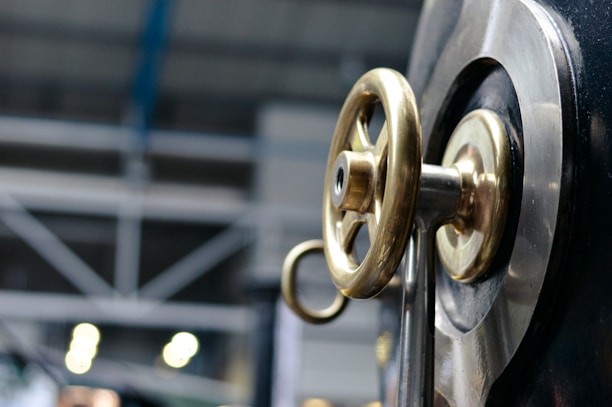 A close-up view of a metallic valve wheel with a brass finish mounted on a large industrial machine. In the background, there are blurred industrial structures and lighting, suggesting a manufacturing or factory setting.