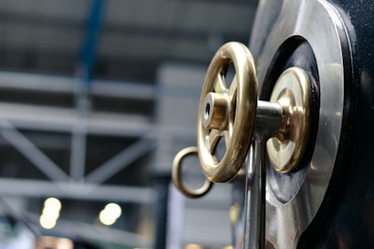 A close-up view of a metallic valve wheel with a brass finish mounted on a large industrial machine. In the background, there are blurred industrial structures and lighting, suggesting a manufacturing or factory setting.