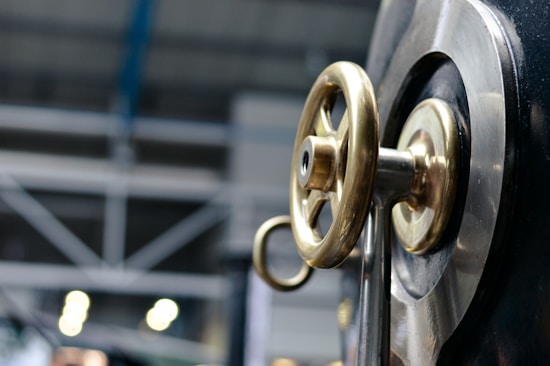 A close-up view of a metallic valve wheel with a brass finish mounted on a large industrial machine. In the background, there are blurred industrial structures and lighting, suggesting a manufacturing or factory setting.