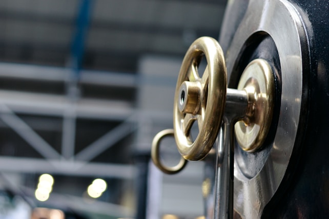 A close-up view of a metallic valve wheel with a brass finish mounted on a large industrial machine. In the background, there are blurred industrial structures and lighting, suggesting a manufacturing or factory setting.