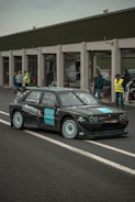 A black rally car with white rims is parked on a race track near a pit lane. The car has various sponsor logos on its body. Several people are standing in the background, some wearing safety vests and observing the vehicle.