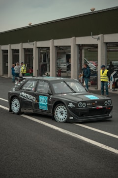 A black rally car with white rims is parked on a race track near a pit lane. The car has various sponsor logos on its body. Several people are standing in the background, some wearing safety vests and observing the vehicle.