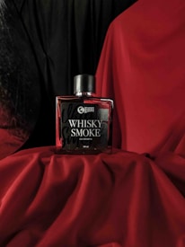 A bottle of Beardo Whisky Smoke eau de parfum is placed on red fabric. The background is a combination of black and red drapery, creating a dramatic and luxurious setting. The bottle has a sleek, rectangular design with a dark cap.