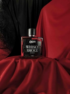 A bottle of Beardo Whisky Smoke eau de parfum is placed on red fabric. The background is a combination of black and red drapery, creating a dramatic and luxurious setting. The bottle has a sleek, rectangular design with a dark cap.