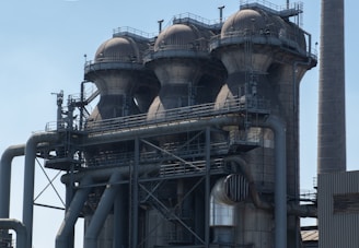 A large industrial structure featuring four cylindrical towers, each topped with a dome-like roof. The structure is made of metal with numerous pipes and platforms connected to it. The surface of the towers is marked by signs of wear, suggesting extensive use. A smokestack or chimney can be seen to the right of the structure against a clear sky.