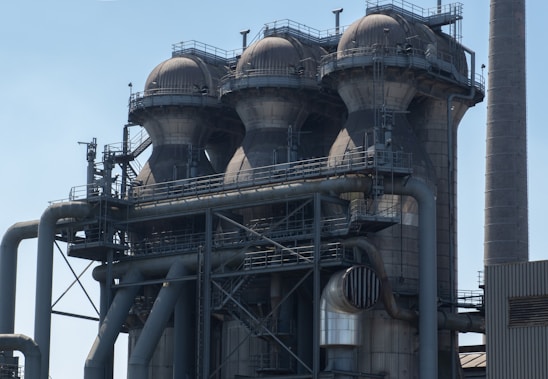 A large industrial structure featuring four cylindrical towers, each topped with a dome-like roof. The structure is made of metal with numerous pipes and platforms connected to it. The surface of the towers is marked by signs of wear, suggesting extensive use. A smokestack or chimney can be seen to the right of the structure against a clear sky.