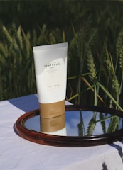 A tube of Madagascar Centella cream is placed on a mirror that reflects the blue sky, set against a backdrop of green wheat fields. The product has a minimalist design with a white body and a brown base, highlighted by natural sunlight.