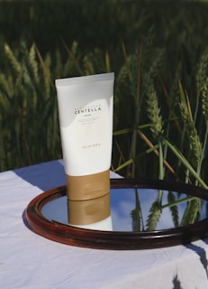 A tube of Madagascar Centella cream is placed on a mirror that reflects the blue sky, set against a backdrop of green wheat fields. The product has a minimalist design with a white body and a brown base, highlighted by natural sunlight.