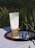 A tube of Madagascar Centella cream is placed on a mirror that reflects the blue sky, set against a backdrop of green wheat fields. The product has a minimalist design with a white body and a brown base, highlighted by natural sunlight.