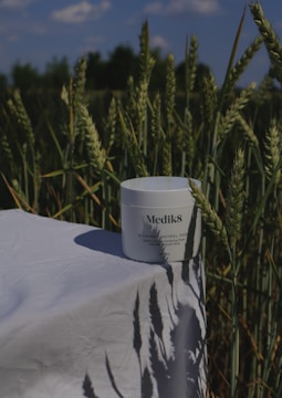 A container labeled 'Medik8 Blemish Control Pads' is placed on a white cloth against a backdrop of a wheat field. The wheat stalks are tall and green, and the sky is partially visible with some clouds. The sunlight casts shadows of the wheat onto the cloth.