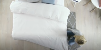 A neatly made bed with white bedding is positioned against wooden flooring. A person with blonde hair is arranging the covers near the foot of the bed. A gray patterned rug is placed partially under the bed. In the corner, there is a white chair with gold legs.