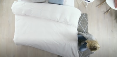 A neatly made bed with white bedding is positioned against wooden flooring. A person with blonde hair is arranging the covers near the foot of the bed. A gray patterned rug is placed partially under the bed. In the corner, there is a white chair with gold legs.
