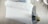A neatly made bed with white bedding is positioned against wooden flooring. A person with blonde hair is arranging the covers near the foot of the bed. A gray patterned rug is placed partially under the bed. In the corner, there is a white chair with gold legs.