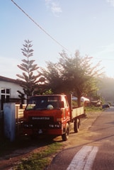 A red Daihatsu truck is parked by the side of a rural road. The scene is set during what appears to be early morning or late afternoon, indicated by the warm sunlight. A tall, coniferous tree and a smaller leafy tree are near the truck, casting shadows on the ground. There are residential buildings in the background, and a distant view of more trees and hills.