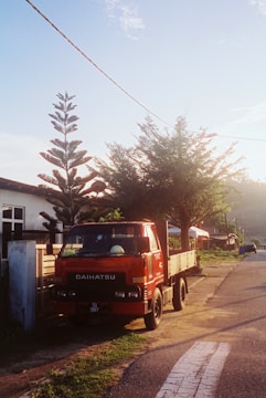 A red Daihatsu truck is parked by the side of a rural road. The scene is set during what appears to be early morning or late afternoon, indicated by the warm sunlight. A tall, coniferous tree and a smaller leafy tree are near the truck, casting shadows on the ground. There are residential buildings in the background, and a distant view of more trees and hills.