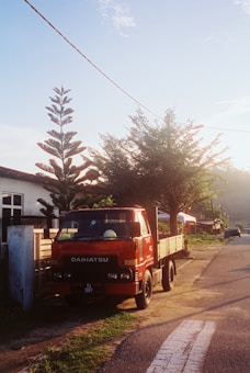 A red Daihatsu truck is parked by the side of a rural road. The scene is set during what appears to be early morning or late afternoon, indicated by the warm sunlight. A tall, coniferous tree and a smaller leafy tree are near the truck, casting shadows on the ground. There are residential buildings in the background, and a distant view of more trees and hills.