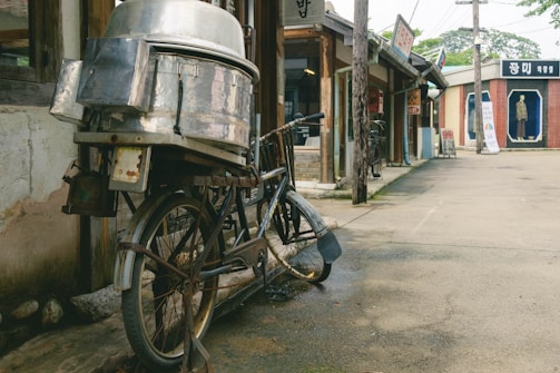 An old bicycle with a large metal container attached to its rear stands against the wall of a vintage building. The street is lined with traditional shops, and a mannequin is displayed in a glass case across the road. Wooden poles and signs hang along the street, evoking a nostalgic, rustic atmosphere.