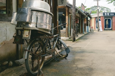 An old bicycle with a large metal container attached to its rear stands against the wall of a vintage building. The street is lined with traditional shops, and a mannequin is displayed in a glass case across the road. Wooden poles and signs hang along the street, evoking a nostalgic, rustic atmosphere.